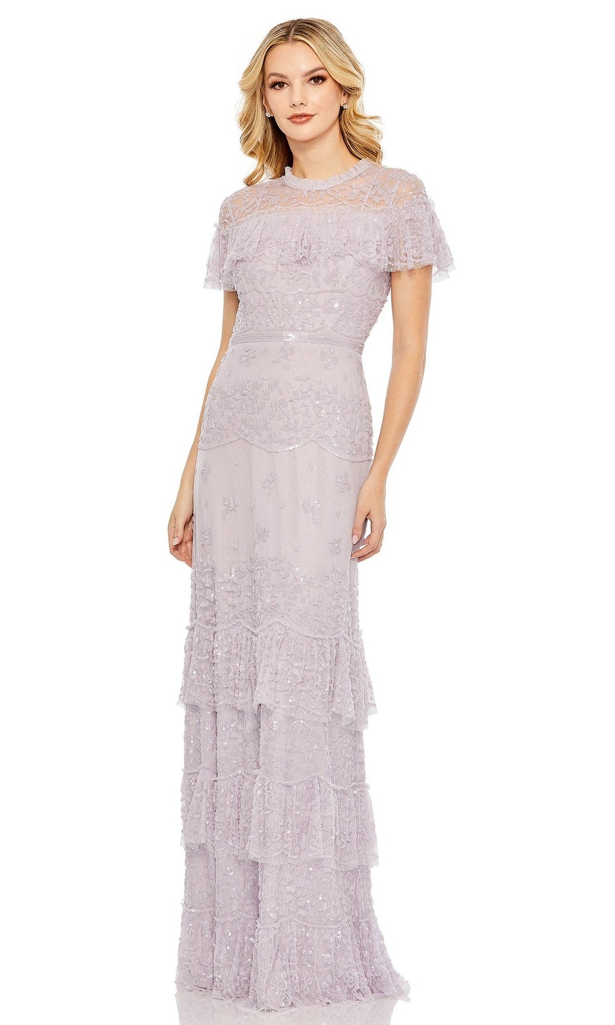 A woman stands wearing the Mac Duggal 9195: a Lilac Long Sequin Formal Dress with short sleeves, floor-length tiered ruffle layers, and sheer details at the shoulders and upper chest.